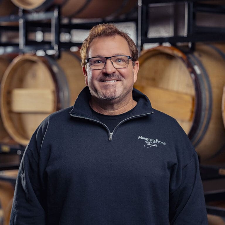Smiling man in glasses and a dark pullover stands in front of wine barrels in a cellar.
