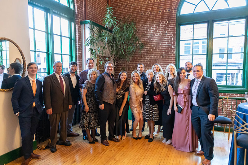 A group of people dressed formally, smiling and posing together in a bright room with large windows and brick walls.