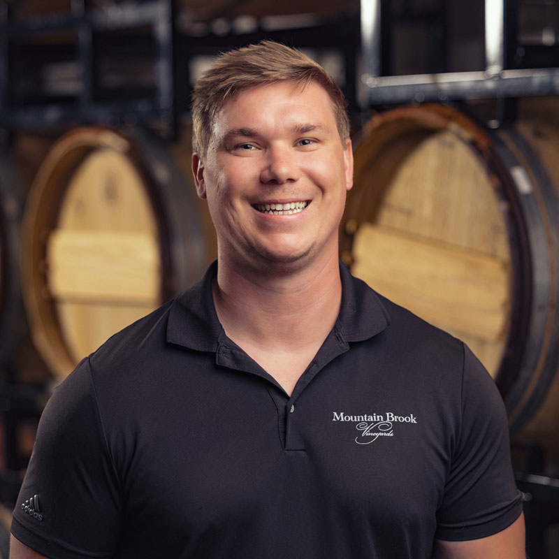 Smiling man in a black polo shirt stands in front of wine barrels in a dimly lit cellar.