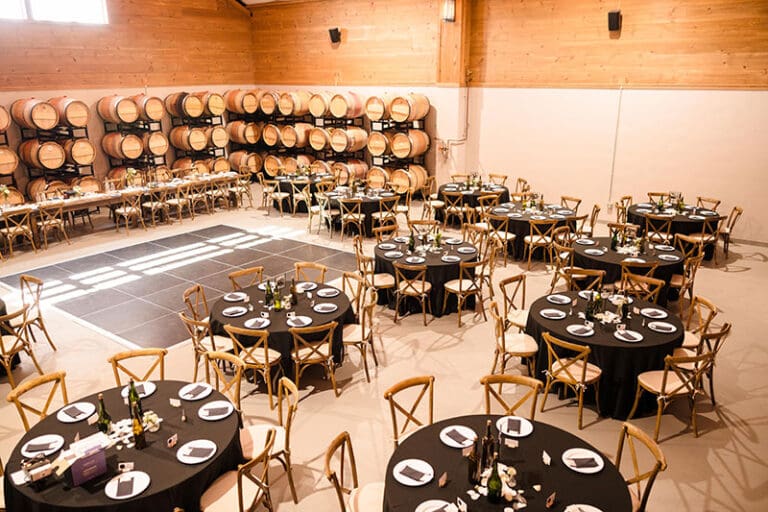 Event hall with round tables set for a meal, wine barrels line the wall, and a dance floor in the center.