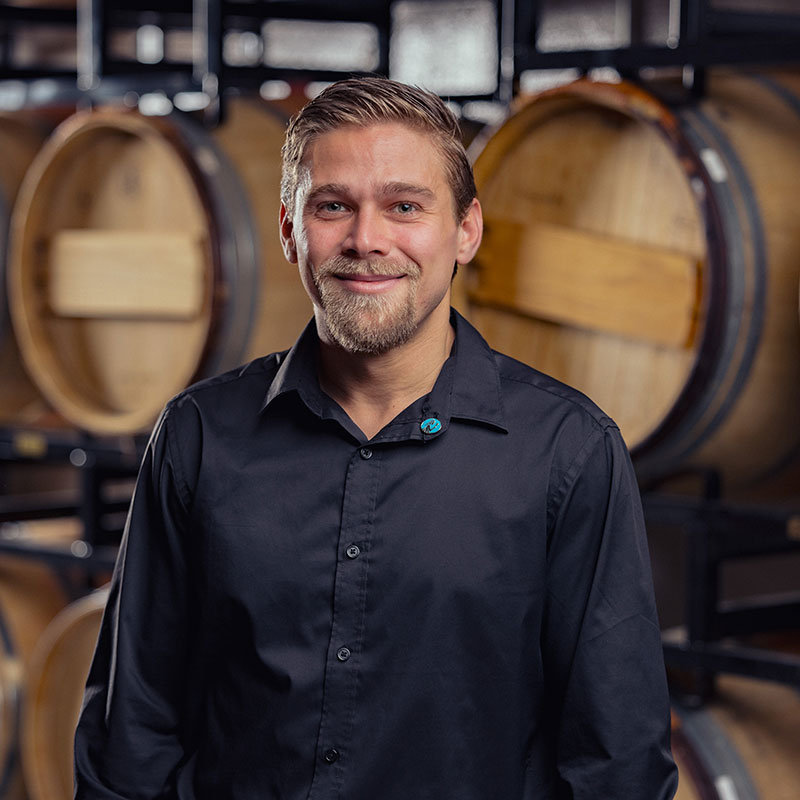 A man in a black shirt smiles in front of stacked wooden barrels at a brewery or winery.