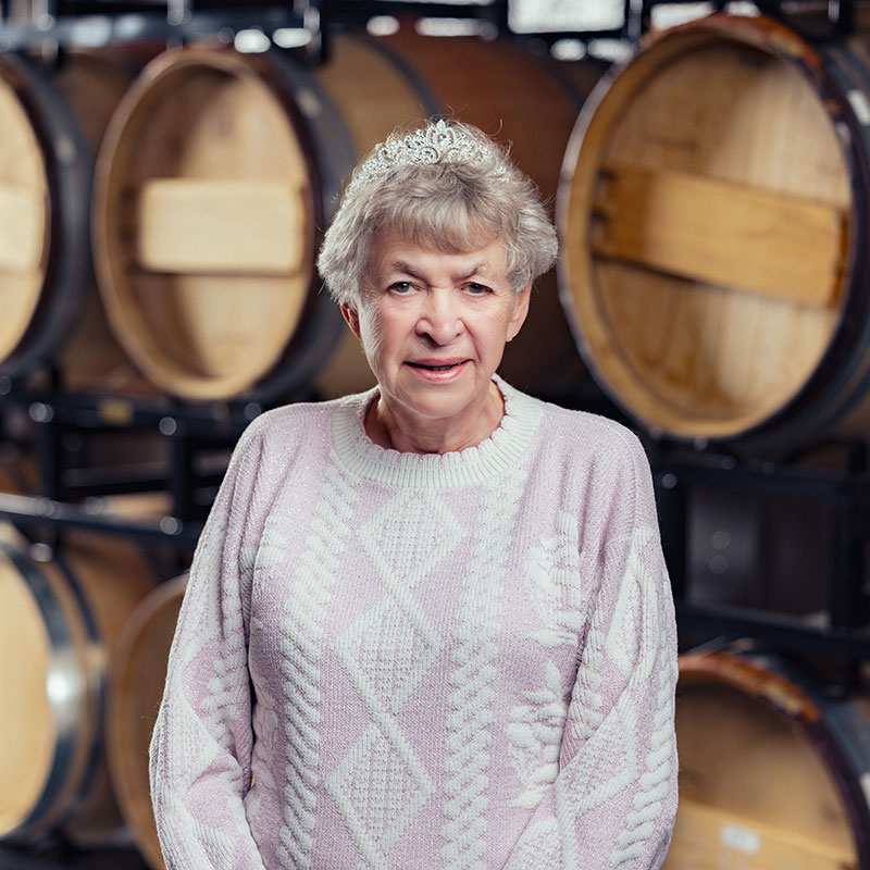 Older woman in a tiara and patterned sweater standing in front of stacked wooden barrels.