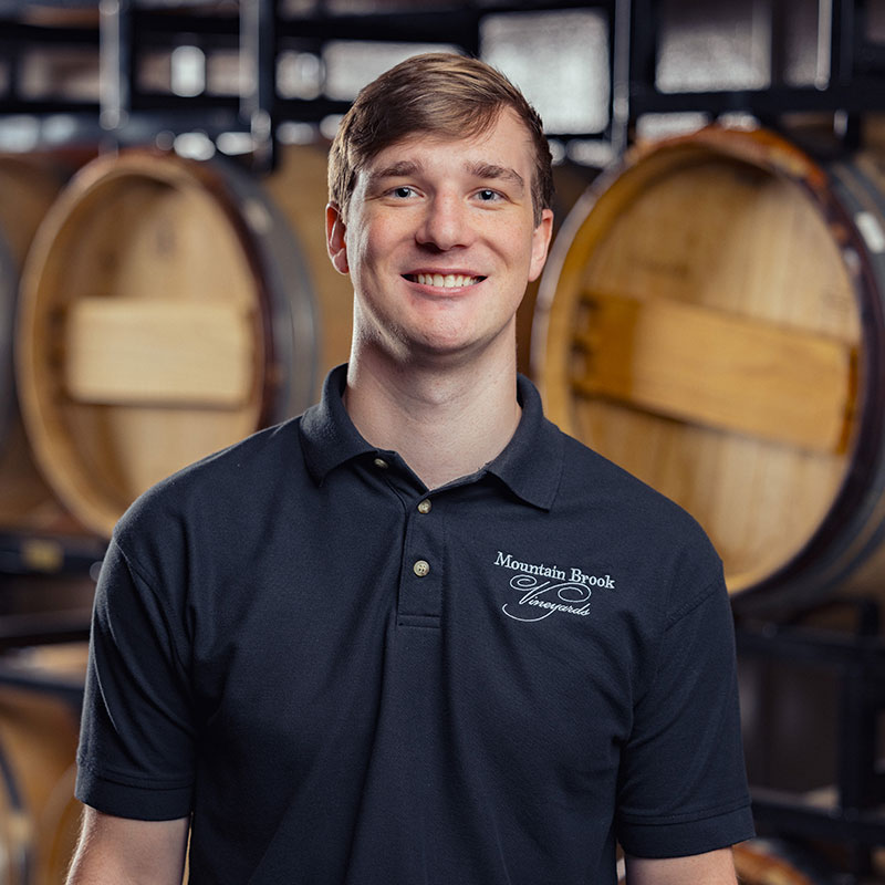 Smiling man in a black Mountain Brook Vineyards polo shirt stands in front of wine barrels.