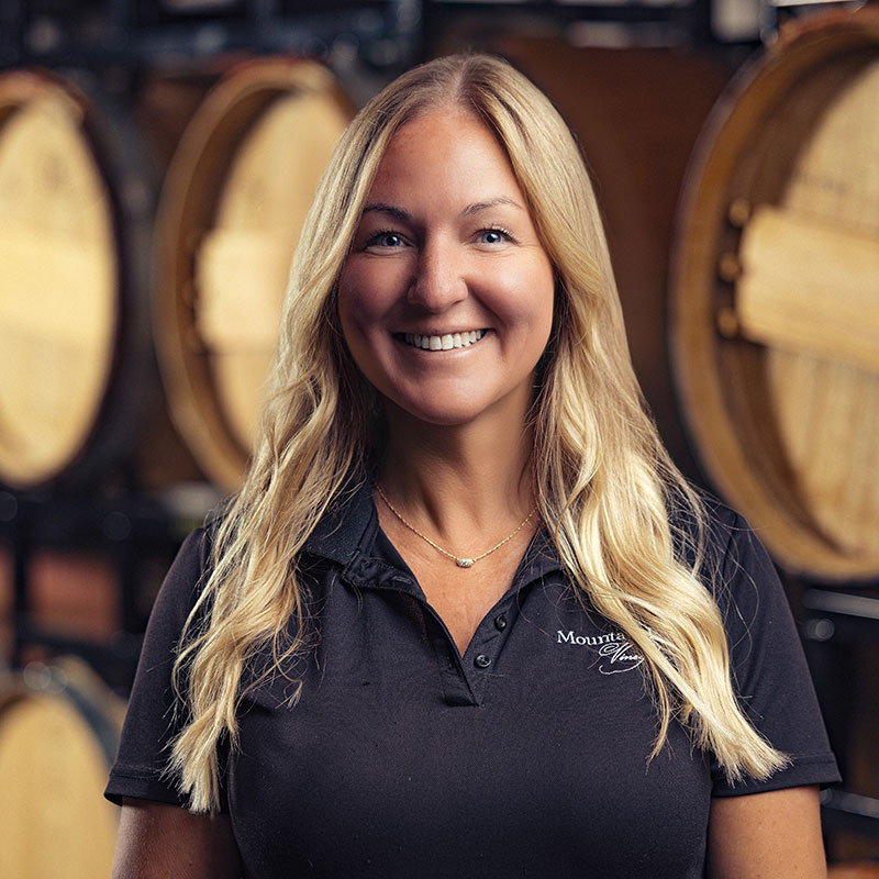 Smiling woman with long blonde hair stands in front of large wooden barrels, wearing a black polo shirt.