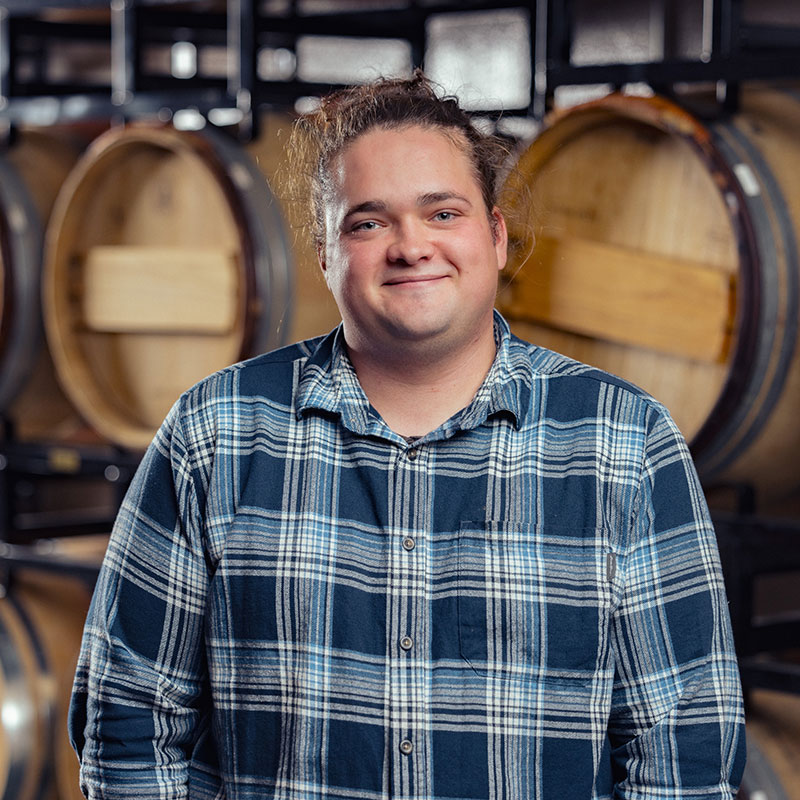 A person in a blue plaid shirt smiles in front of stacked wooden barrels in a brewery or winery.