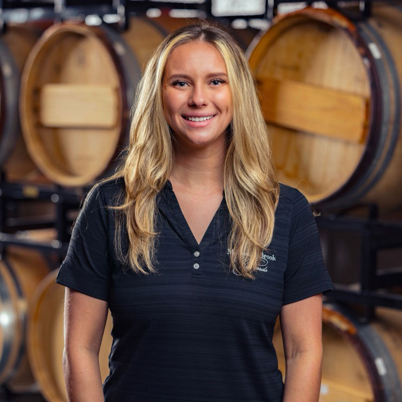 Smiling woman with long blonde hair stands in front of stacked wooden barrels in a winery.