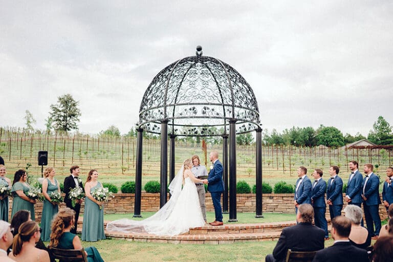 Bride and groom exchange vows under a decorative gazebo, surrounded by their wedding party and guests outdoors.