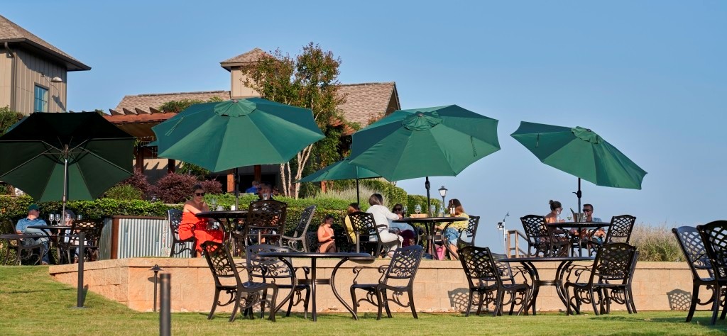 People sitting at outdoor tables under green umbrellas on a sunny day near a building with a sloped roof.