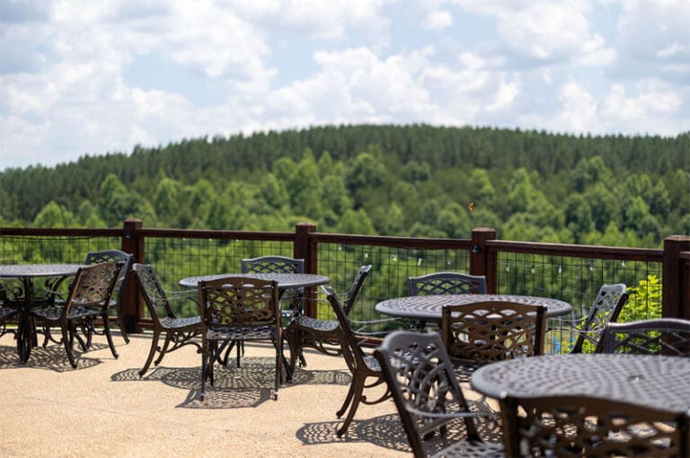 Outdoor patio with empty metal tables and chairs overlooking a lush green forest and cloudy sky.