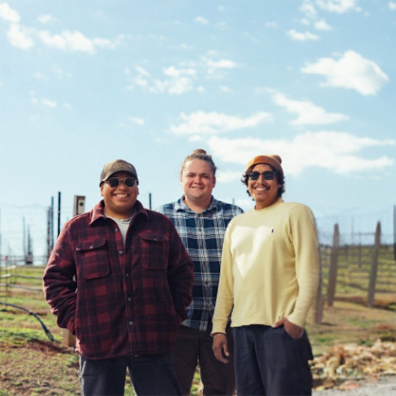 Three men smiling and standing outdoors in front of a vineyard under a blue sky with scattered clouds.