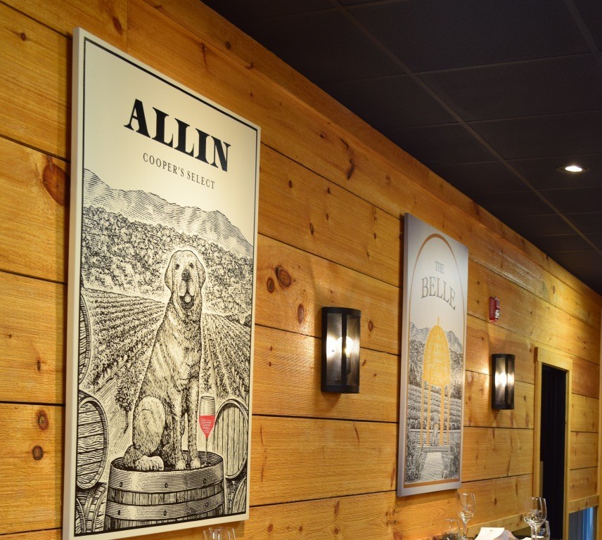 Framed dog illustration and vineyard print on a wooden wall in a warmly lit restaurant or tasting room.
