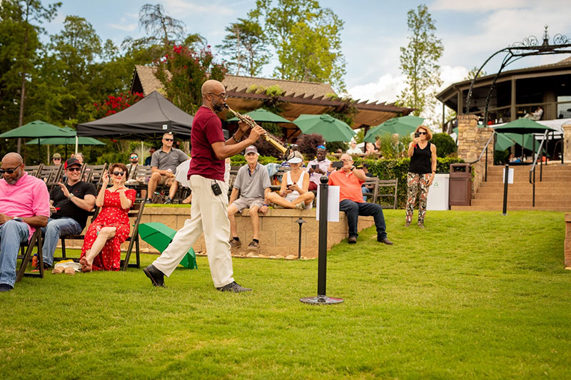 A man plays saxophone outdoors while people sit on lawn chairs and watch on a sunny day.