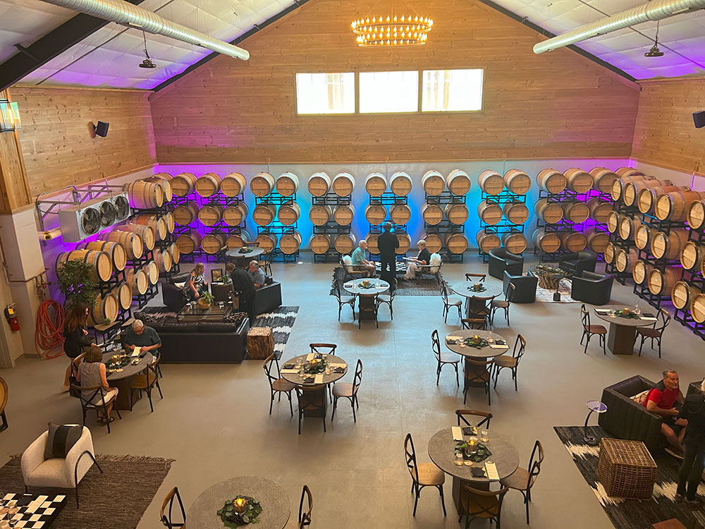 Spacious winery tasting room with wine barrels on walls, round tables, and people seated and chatting.