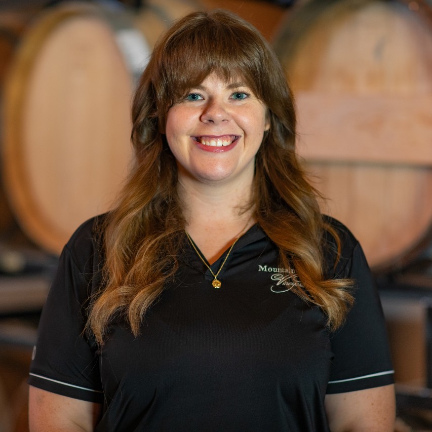 Smiling woman with long brown hair standing in front of large wooden barrels, wearing a black shirt.